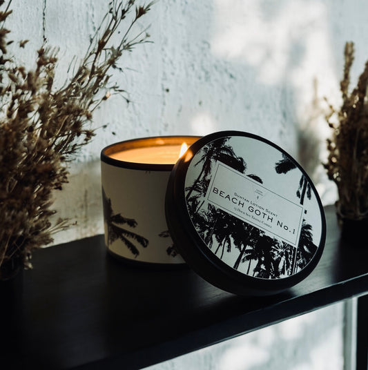 Lit “Beach Goth No. 1” candle with a black-and-white palm tree design sits on a black shelf between dried floral arrangements. The label highlights a suntan lotion scent, evoking summer nostalgia with a dark, moody twist. Perfect for beach lovers with a gothic edge—adds ambiance and personality to any space