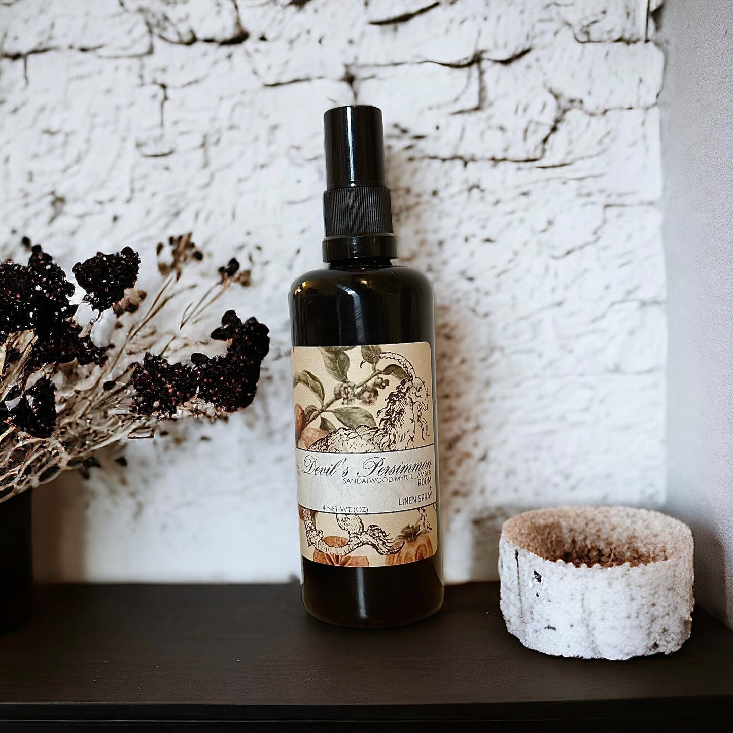 A black spray bottle labeled “Devil’s Persimmon” with a vintage-style design featuring a goat and botanical elements. Scent notes include sandalwood, myrtle, and amber. The bottle rests against a textured white background with dried florals nearby.