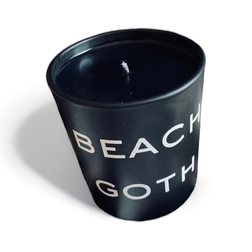 Black candle labeled “BEACH GOTH” in bold white letters, housed in a matte black glass jar. Ideal for gothic home decor, alternative beach vibes, and unique candle lovers.