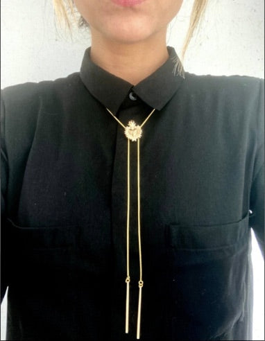 A gold plated bolo tie with a detailed centerpiece featuring a sacred heart design is worn over a black button-up shirt. The tie has two long, delicate gold plated chains ending in slim metal bars
