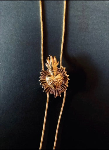 Closeup of gold plated bolo tie with a detailed centerpiece featuring a radiant heart design with black background.The tie has two long, delicate gold plated chains ending in slim metal bar