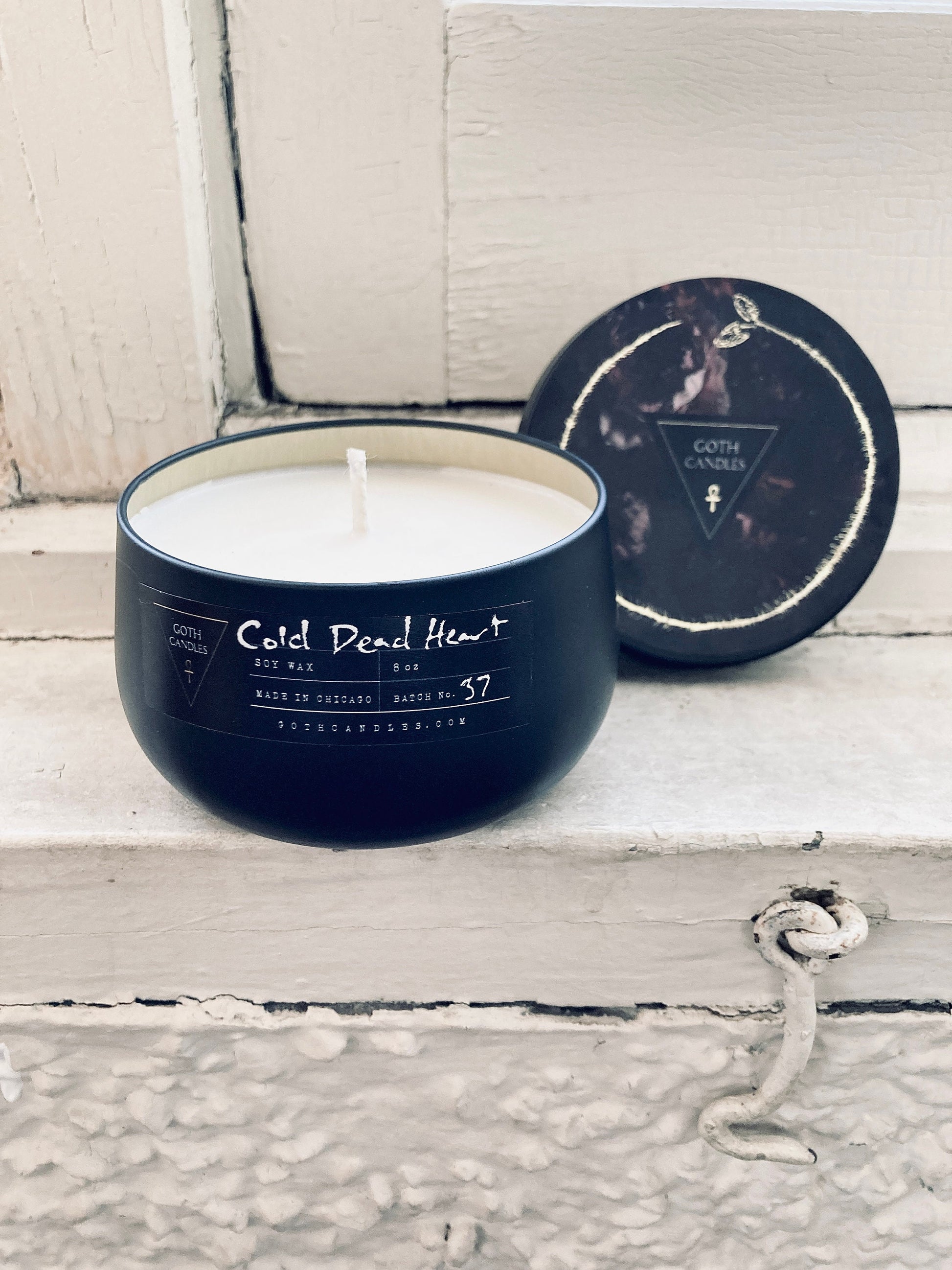A black soy wax candle labeled “Cold Dead Heart” in gothic-style typography. It comes in a dark container with a matching lid featuring a minimalist triangle logo. The candle has a single white wick and a moody design perfect for gothic aesthetics.