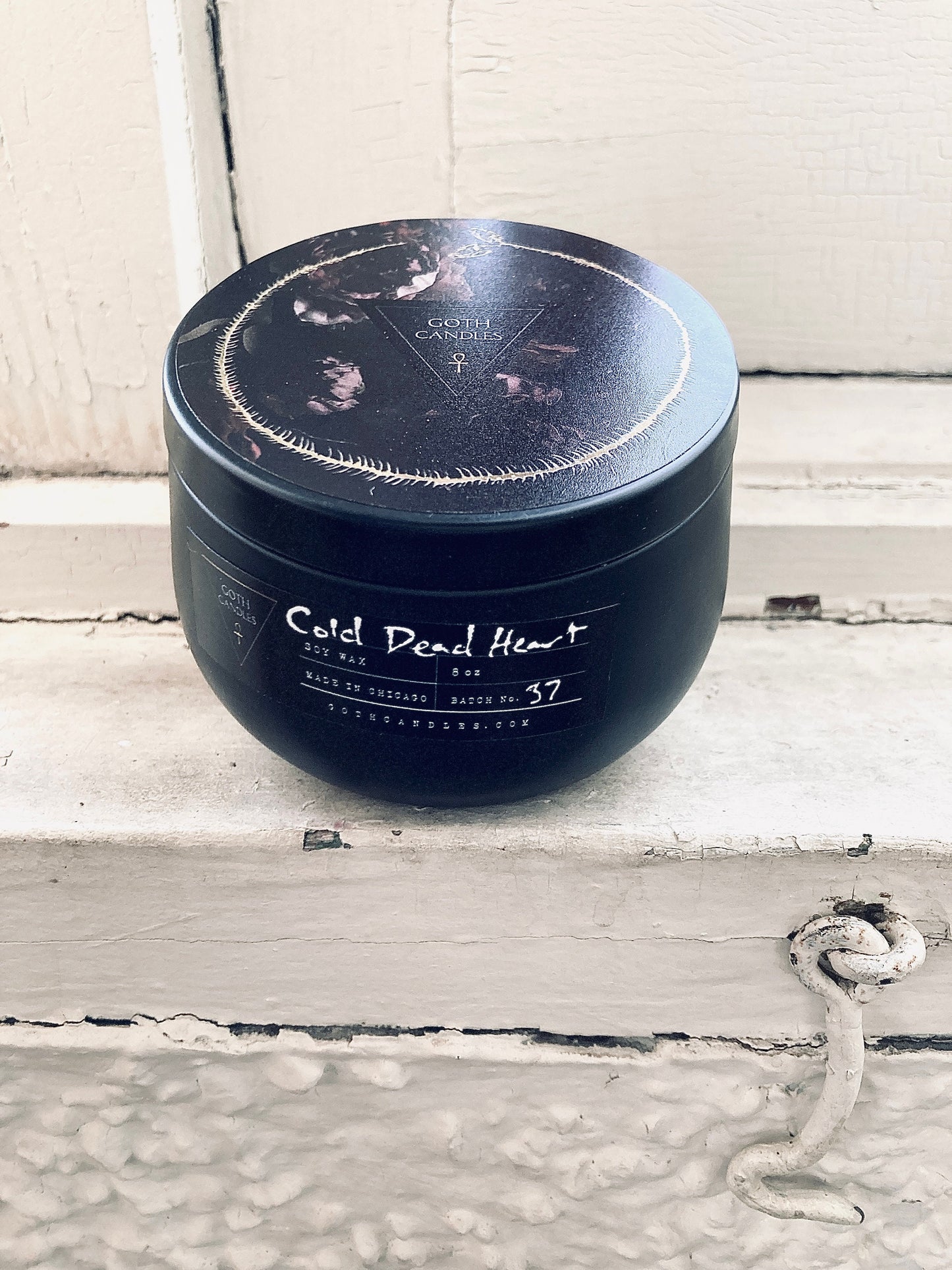 A black soy wax candle labeled “Cold Dead Heart” in gothic-style typography. It comes in a dark container with a matching lid featuring a minimalist triangle logo. The candle has a single white wick and a moody design perfect for gothic aesthetics.