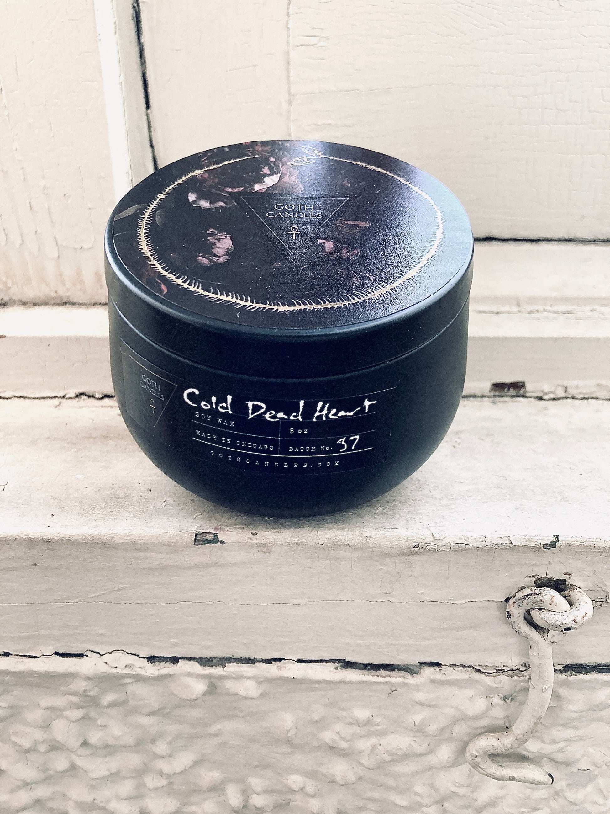 A black soy wax candle labeled “Cold Dead Heart” in gothic-style typography. It comes in a dark container with a matching lid featuring a minimalist triangle logo. The candle has a single white wick and a moody design perfect for gothic aesthetics.