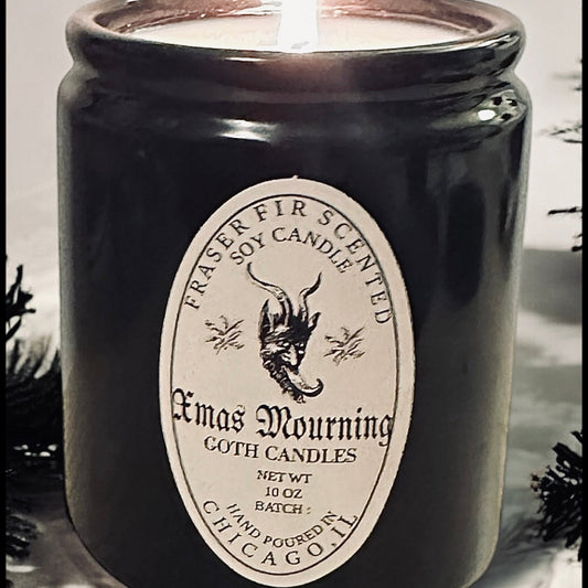 Black candle labeled 'Xmas Mourning' with a white label featuring text and graphics, surrounded by greenery.