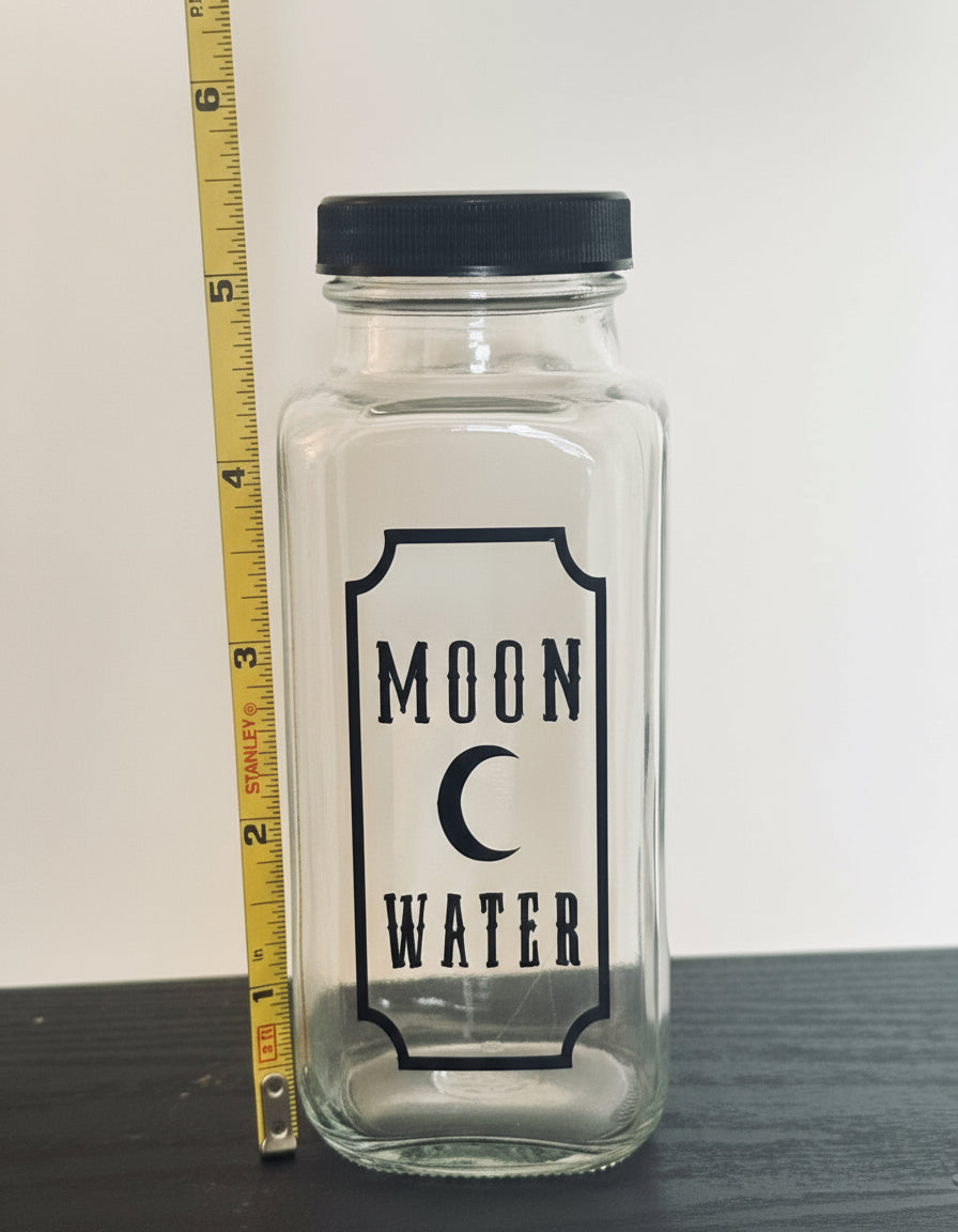 Clear glass jar labeled 'Moon Water' with a black lid, next to a measuring tape for scale.
