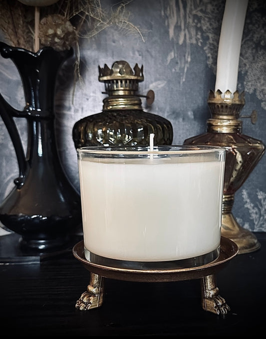A white candle in a clear glass container sits on an ornate bronze stand with clawed feet. Behind it, vintage decor including a black vase, a green glass oil lamp with a crown-shaped top, and a brass candlestick create a gothic, antique atmosphere