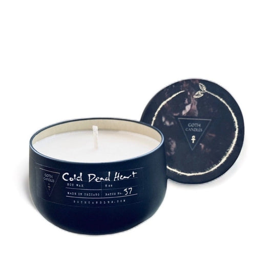 A black soy wax candle labeled “Cold Dead Heart” in gothic-style typography. It comes in a dark container with a matching lid featuring a minimalist triangle logo. The candle has a single white wick and a moody design perfect for gothic aesthetics.