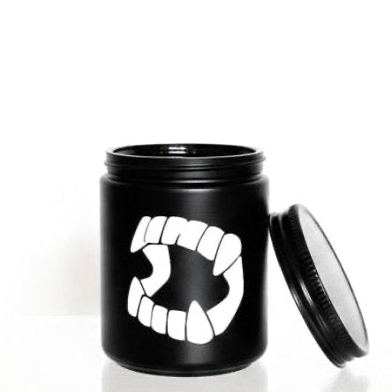 A small black jar with a matching lid leaning against it on a white reflective surface. The jar has a bold white graphic of a mouth with sharp teeth on the front.
