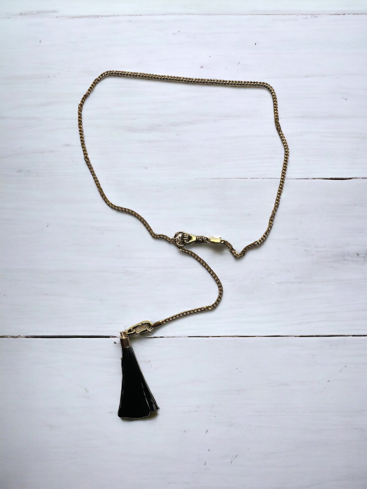 An adjustable lariat necklace with a gold-plated chain and a black tassel charm, displayed against a white wooden background.