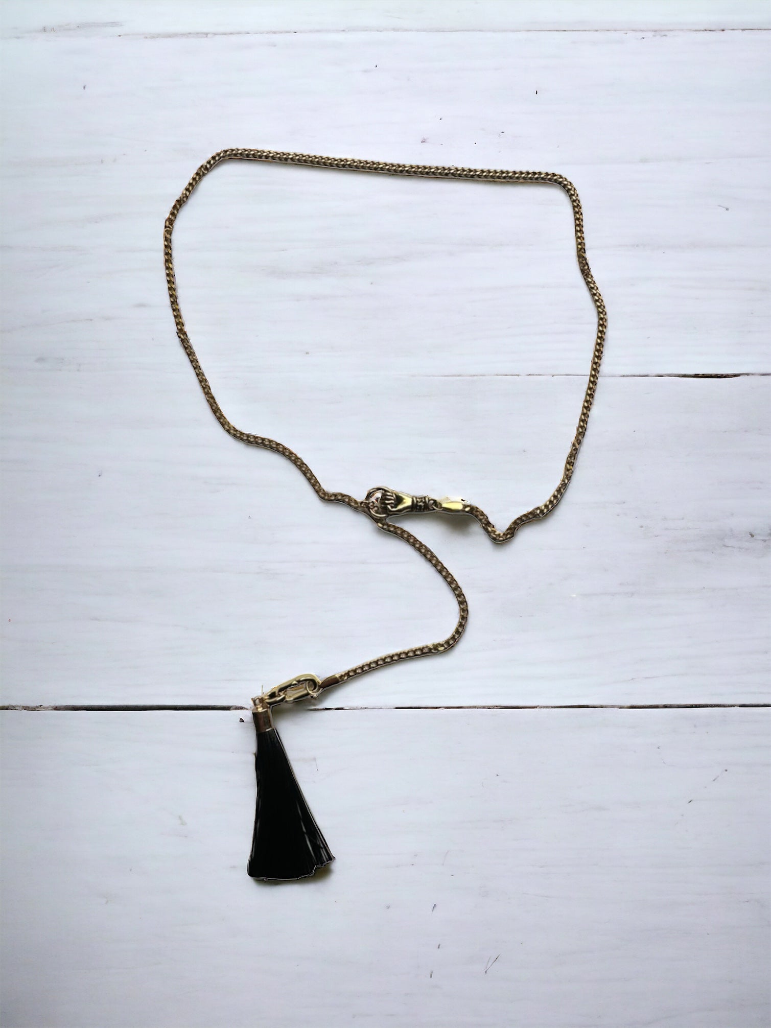 An adjustable lariat necklace with a gold-plated chain and a black tassel charm, displayed against a white wooden background.