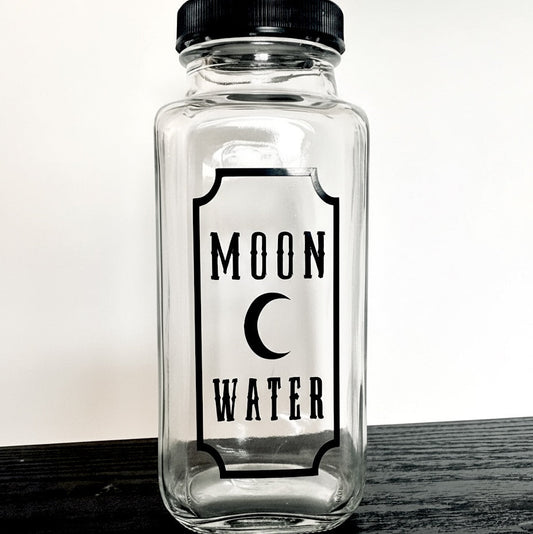 Clear glass jar with black lid and 'Moon Water' label on a wooden surface.
