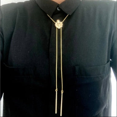 A gold plated bolo tie with a detailed centerpiece featuring a sacred heart design is worn over a black button-up shirt. The tie has two long, delicate gold plated chains ending in slim metal bars